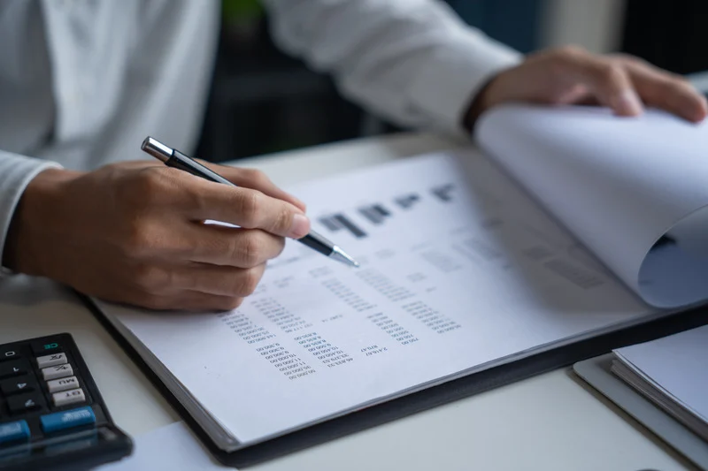 Person writes on financial documents with a pen, a calculator visible nearby on a desk.