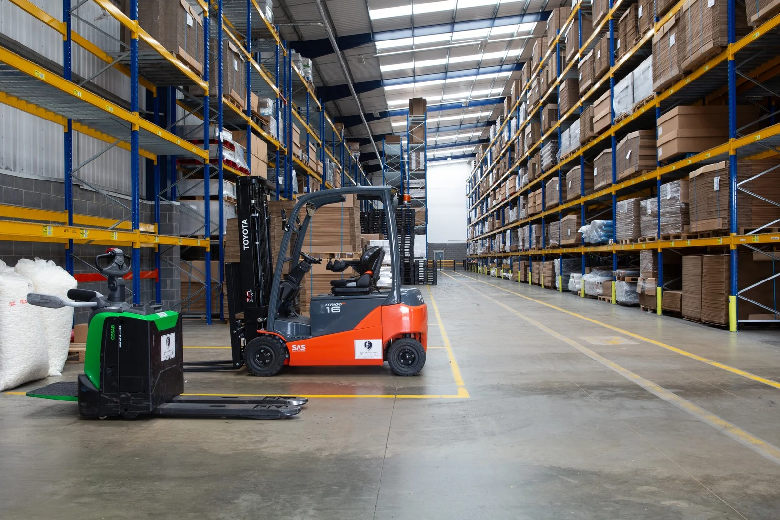 Orange forklift in a large warehouse aisle, surrounded by tall blue and yellow pallet racks filled with boxes.