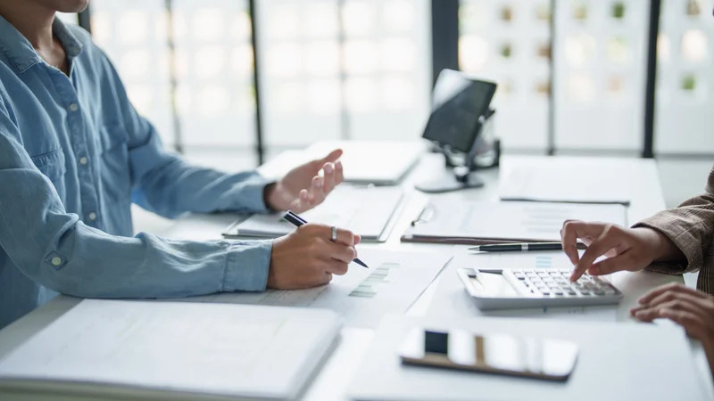 s Two people at a bright office desk reviewing documents; one writes with a pen while the other uses a calculator.