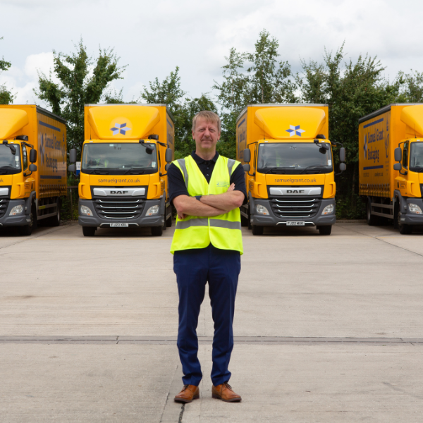 sgp ag Man in a high-visibility vest standing with arms crossed in front of yellow delivery trucks behind him in a parking lot.