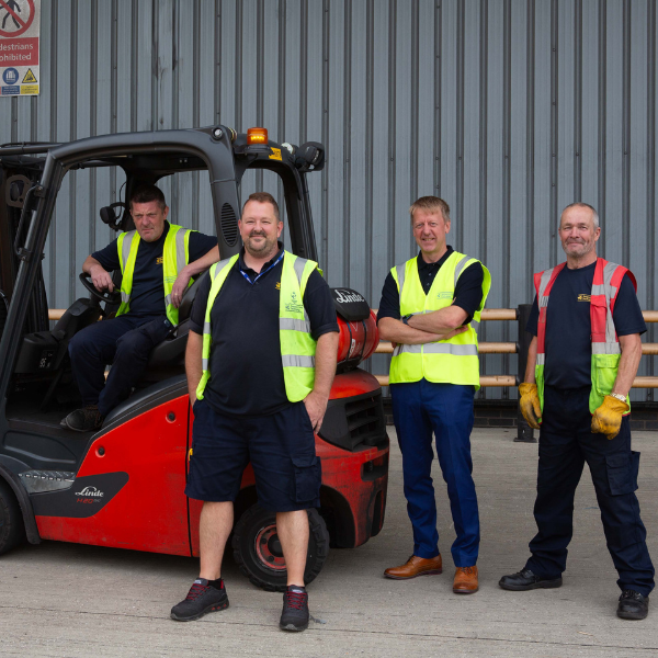 sgp Four workers in high-visibility vests stand beside a red forklift outside a corrugated metal building; one sits in the driver seat.