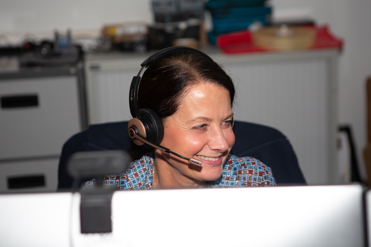 sgp2 Woman wearing a headset and smiling at a desk, likely engaged in a call or video chat in an office setting.