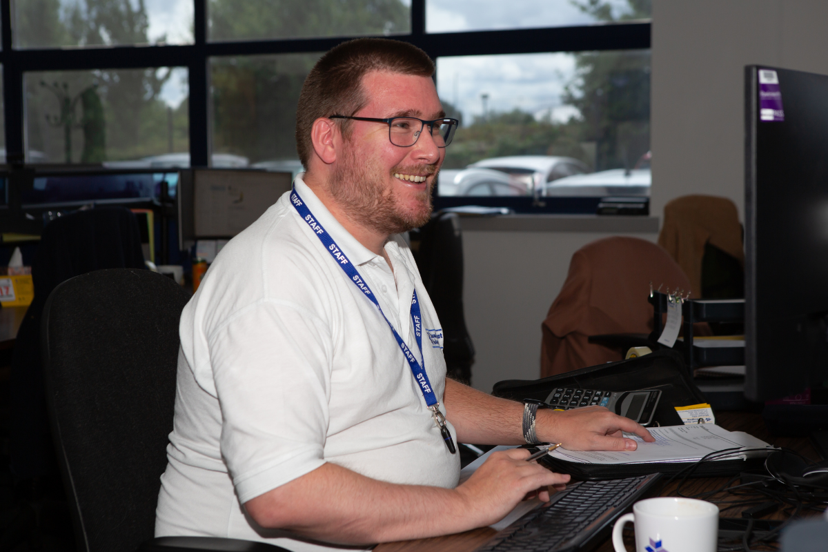 sgp3 Smiling man wearing glasses and a blue 'STAFF' lanyard sits at a desk in an office.