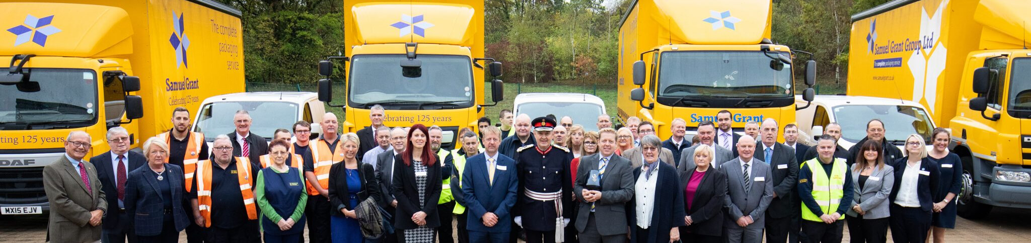 why us img Group of company employees and officials posing in front of bright yellow Samuel Grant delivery trucks at a 125-year celebration event.