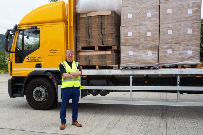 width_800 Delivery driver in a neon safety vest stands with arms crossed beside a yellow truck loaded with pallets and cargo on a trailer.